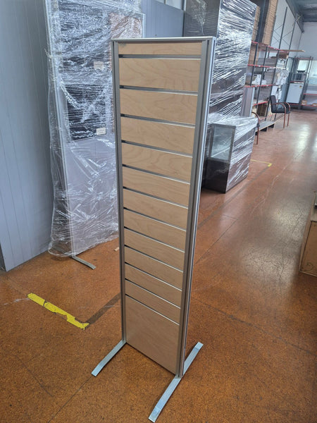 A tall two sided slatwall display panel with a light wood grain finish and aluminium framing, shown standing on a warehouse floor next to other wrapped furniture pieces.