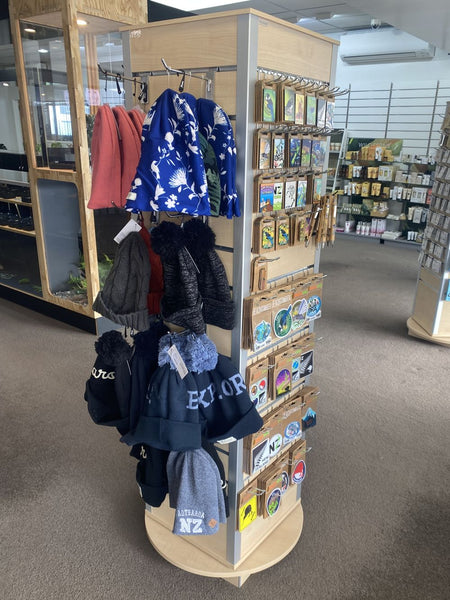 A rotating retail display stand in a store showing children's clothing and small packaged accessories. The stand holds several jackets and hoodies in blue, black, gray, and coral, along with many small cards or packets attached to the side panels.