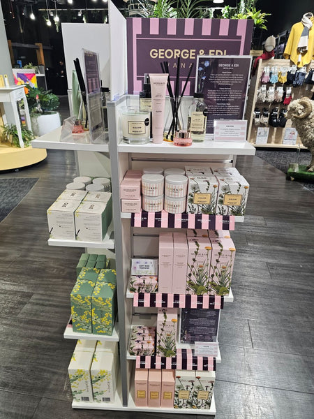 A retail display stand filled with George & Ed's home fragrance products, including candles, diffusers, and boxed items, set in a trade expo with dark wood floors and other merchandise in the background.