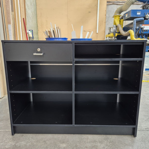 back view of a black cashier desk with open adjustable shelves and a lockable drawer with a metal handle, photographed in a warehouse setting.