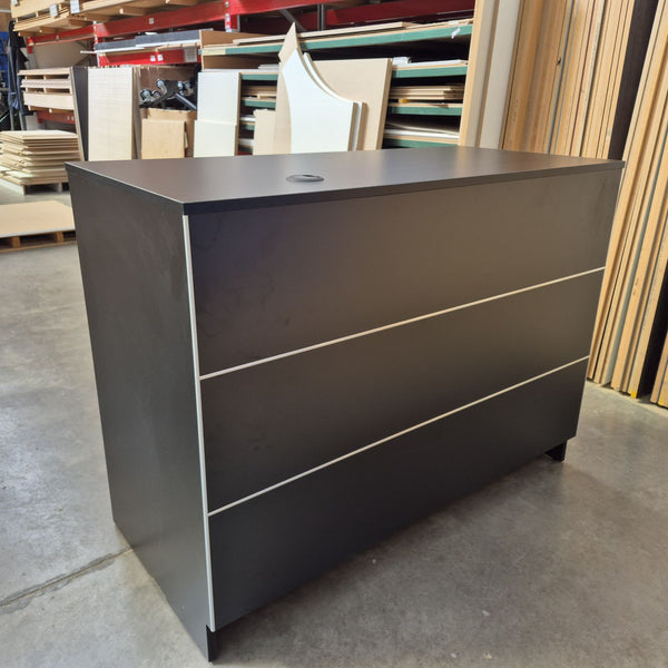 Front view of a black Cashier Desk with thin silver detailing, photographed in a warehouse setting.