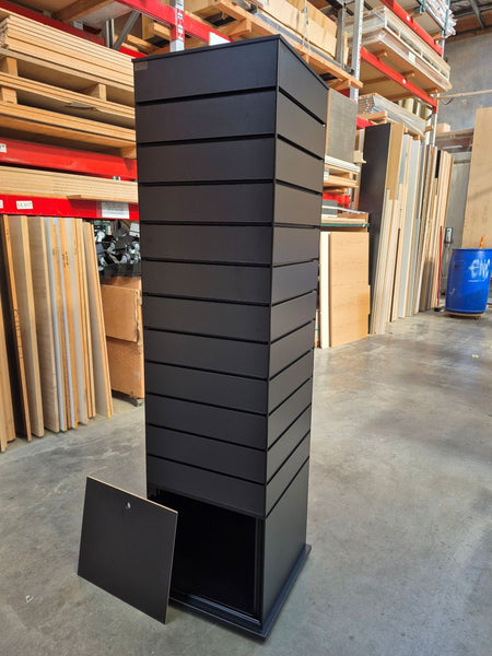 A tall, black slimline slatwall four sided retail display tower with an open storage cupboard in its base, photographed in the Plankwall warehouse in front of wood panels and materials.