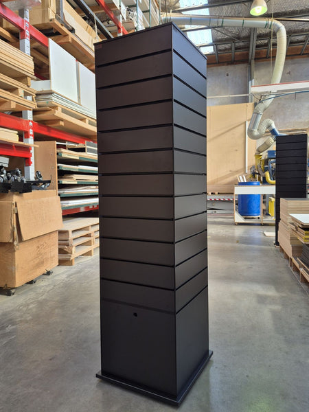 A front view of a tall, black slimline slatwall four sided retail display tower photographed in the Plankwall warehouse in front of wood panels and materials.