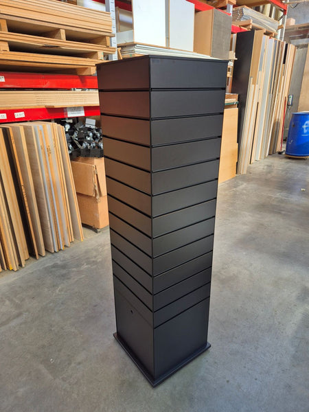 A side view of a tall, black slimline slatwall four sided retail display tower photographed in the Plankwall warehouse in front of wood panels and materials.