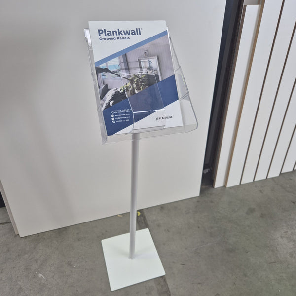 A clear acrylic brochure holder on a white metal floor stand, displaying a product brochure in an indoor showroom setting.