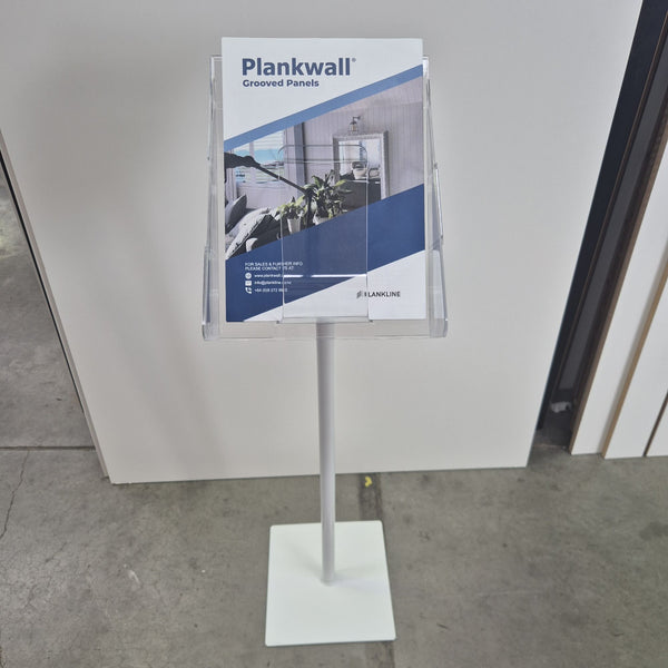 A white freestanding magazine or brochure stand with a clear angled holder on top, shown standing front on  on a concrete floor against a white wall.