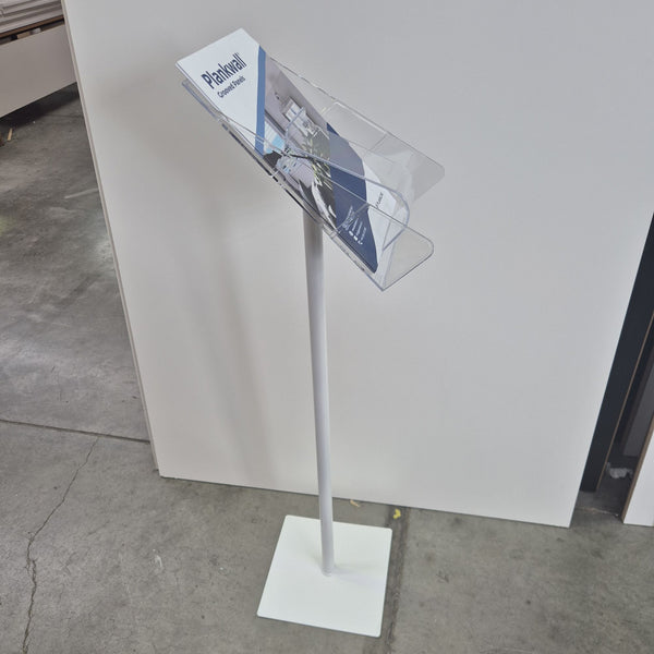 A white freestanding magazine or brochure stand with a clear angled holder on top, shown standing on a concrete floor against a white wall.