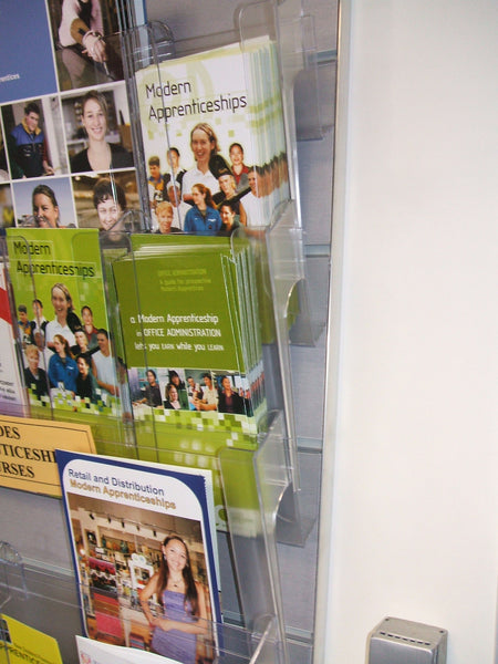 Close up of brochures on display in a Calext clear wall-mounted modular print display system  on a Plankwall panel affixed to the wall.