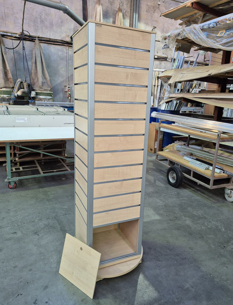 A tall slatwall four sided retail display  unit with a light maple wood finish and aluminium  trim with a storage cabinet in base, photographed in a warehouse or workshop setting.
