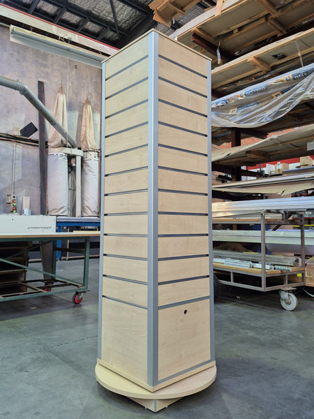 A tall slatwall four sided retail display  unit with a light maple wood finish and aluminium  trim, photographed in a warehouse or workshop setting.