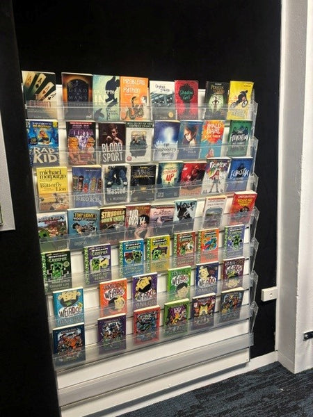 A wall-mounted clear acrylic slatwall calext book display rack filled with colorful book covers, shown in an indoor room with dark walls