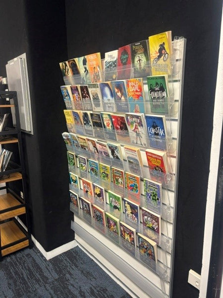 A wall-mounted clear acrylic slatwall calext book display rack filled with colorful book covers, shown in an indoor room with dark walls and a carpeted floor.