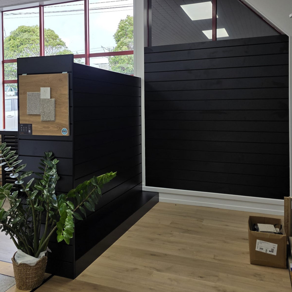 A modern interior space with a prominent black slatted display wall and matching black cabinet, set on light wood flooring with large windows, ceiling lights, and a potted plant.