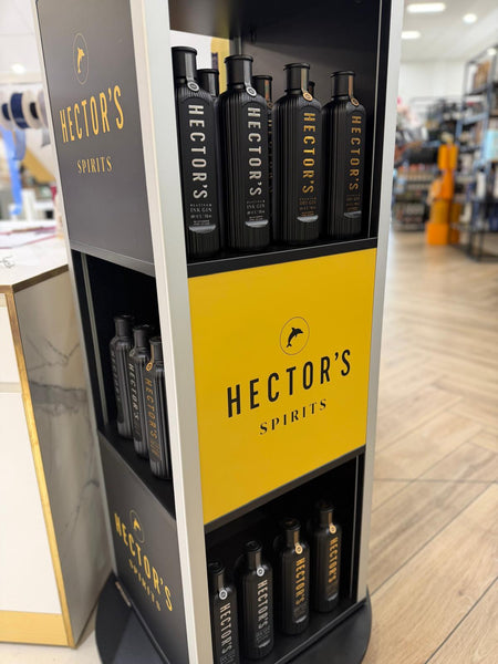 A retail display stand for Hector's Spirits, featuring black bottles with gold lettering on dark gray and bright yellow panels, set in a store environment with wooden floors and blurred background shelves.