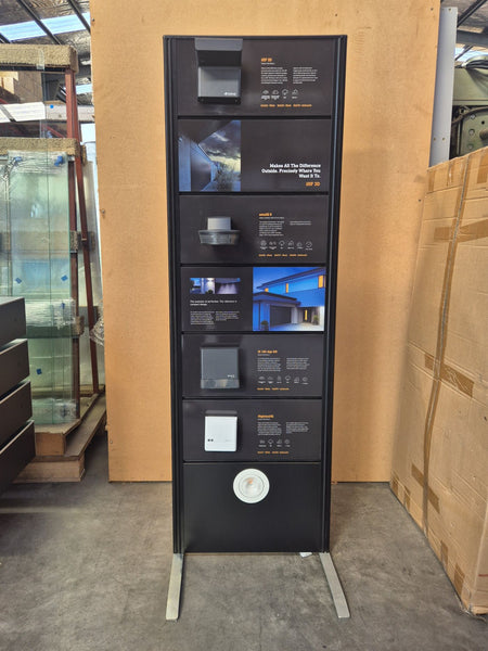 A tall black freestanding display panel with light samples photographed in a warehouse-like indoor setting with cardboard boxes and glass tanks visible around it.