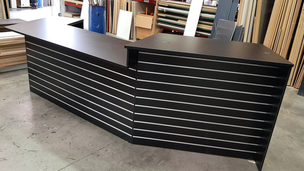 Plankwall L-shaped retail counter and reception desk front with slatwall display space in a warehouse setting with various items in the background