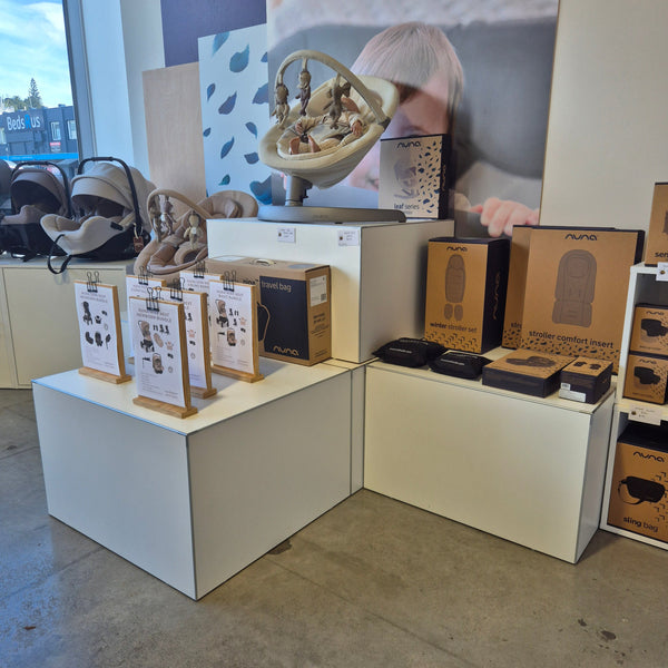 Instore product merchandising of baby accessories on white display cubes in a retail store with a cityscape view outside.