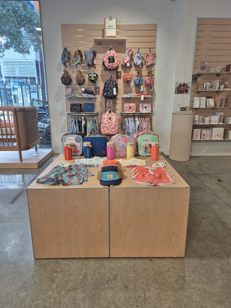 Display of various products on a set of blonde wood display cubes in a retail store selling baby goods with a matching slatwall wall panel hung with baby clothes and a cityscape background