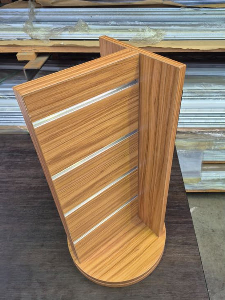 A light brown woodgrain slatwall four sided counter top display stand with a slatted front panel and a circular base, photographed in a workshop area with stacked materials in the background.
