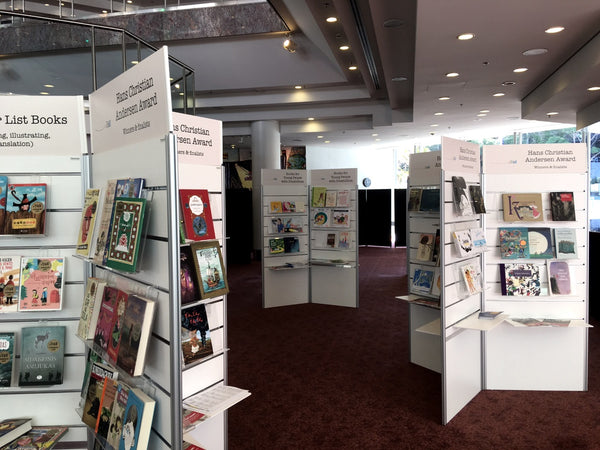 Book display area with books displayed on  clear sloping display shelves mounted on Plankwall slatwall panels in an exhibition space
