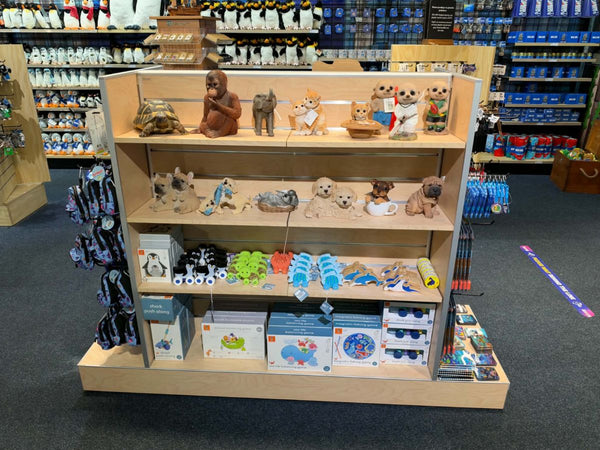 A wooden retail display natural wood gondola with shelves filled with assorted plush toys, figurines, and boxed merchandise, photographed inside a retail store. The shelf has multiple tiers and is surrounded by store fixtures and shelving in the background.