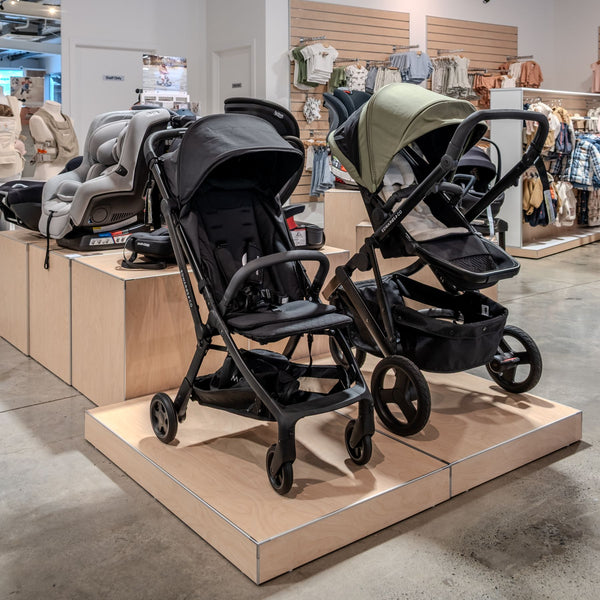 Two black strollers on display in a store setting with baby products in the background.