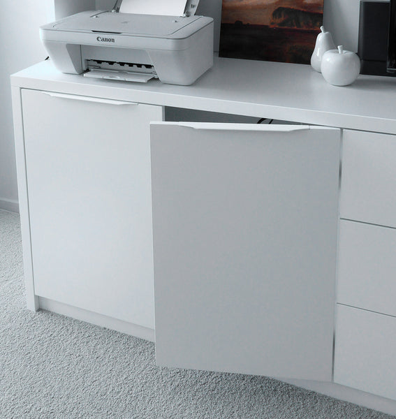 A white built-in cabinet door with a slim horizontal handle, shown slightly open on a matching white desk or dresser. A Canon printer sits on top, with a framed picture and white decorative objects in the background, and light gray carpet on the floor.