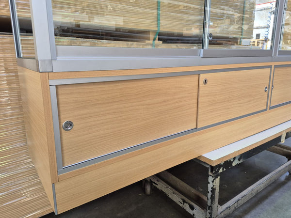 A light oak wood display cabinet with silver metal trim and sliding doors using the Plankwall door track and filler, shown in a warehouse setting.