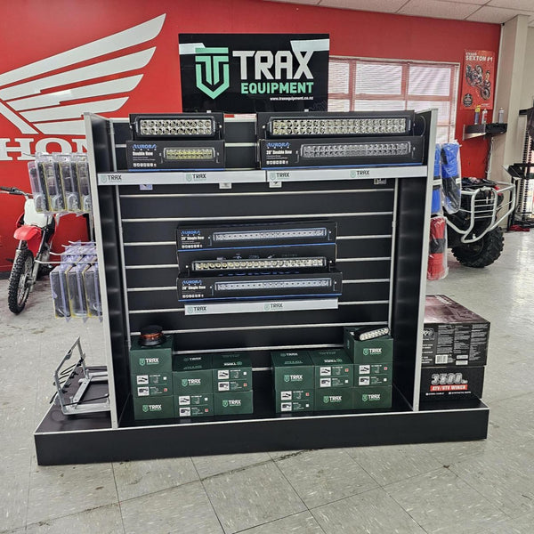 A black and silver TRAX Equipment display stand showcasing off-road light bars and green TRAX batteries in a showroom setting.