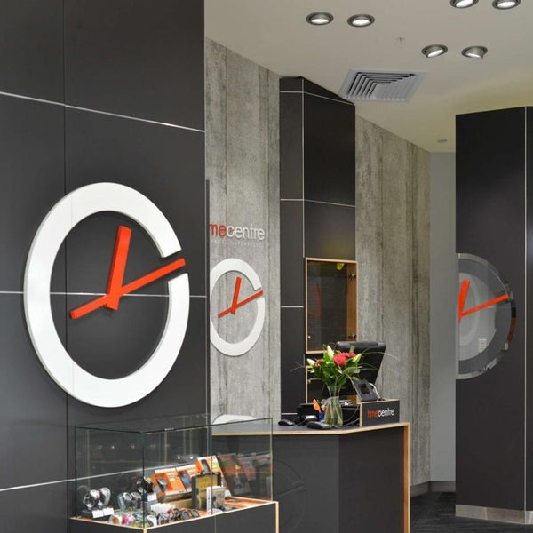 Time Centre shop interior with three large decorative wall clocks with white circular frames and bright orange hands on black walls with MCBUT butt join feature joinery. A reception desk with a glass display case and a vase of flowers is visible in the foreground.