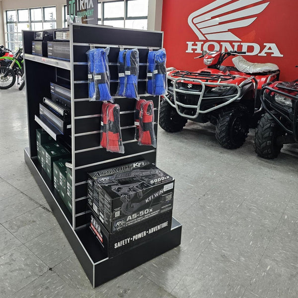 A black retail display stand in a Honda showroom, holding boxed products and blue/red packaged items on a slatwall. Red Honda ATVs are visible in the background along with a large Honda logo on a red wall.