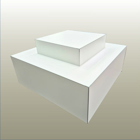 A white two-tiered display retail plinth with a square base and a smaller square box stacked on top, shown against a neutral light gray gradient background.