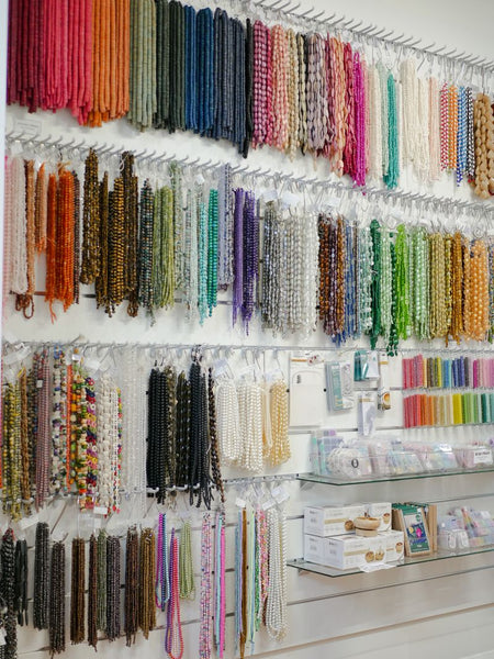 A retail display wall filled with numerous strands of beaded necklaces and bracelets in many colors, hanging on white hooks against a 150 wide slatwall panel, with glass shelves holding packaged items below.
