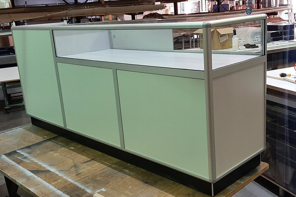 A white retail counter unit with a large glass display counter top in a factory setting - front view