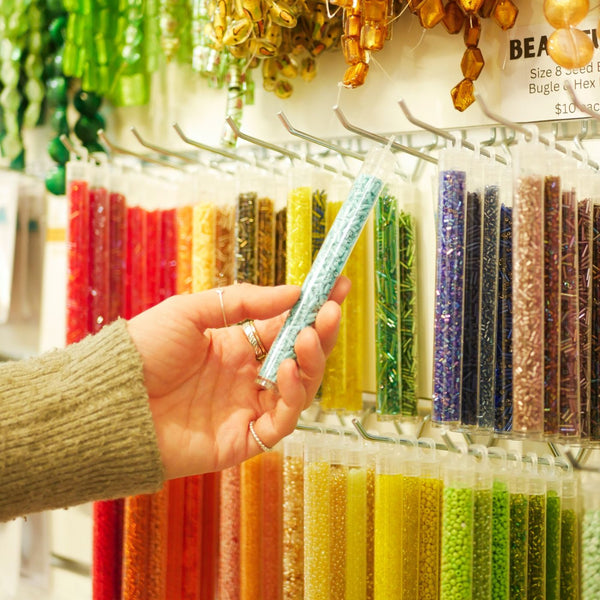 A hand wearing a beige knit sweater holds a clear plastic tube filled with light blue glitter beads in front of a display wall of colorful craft beads hung on slatwall with Plankwall light duty single prongs.
