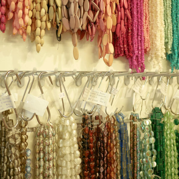 A display of colorful beaded necklaces and bracelets hanging from polycarbonate hang sell hooks on a wall, with many strands in pink, red, green, blue, white, and beige tones.