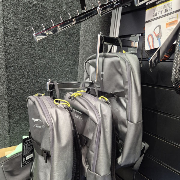 Gray backpacks hanging on a cranked arm display bracket rack with a black slatwall in the background