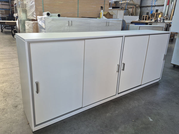 A Plankwall long white MDF storage cabinet with four sliding doors and slim silver trim and handles, photographed in a warehouse or showroom setting.