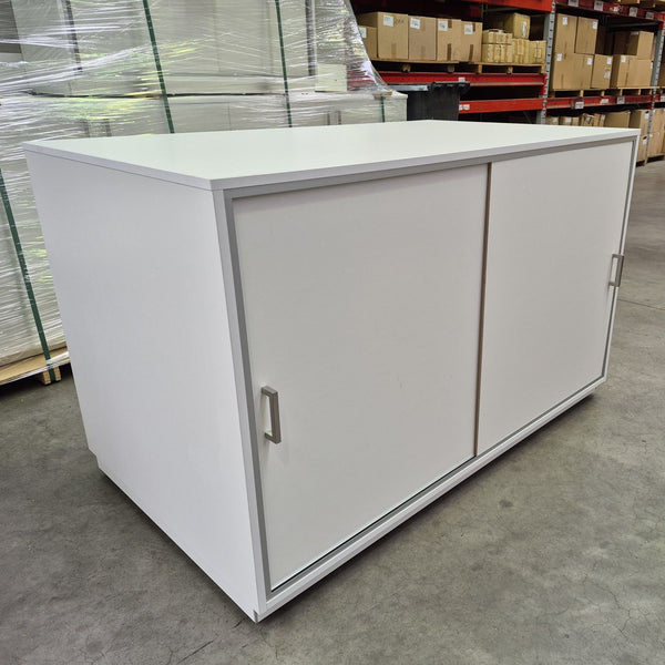 A Plankwall white MDF storage cabinet with two sliding doors and silver handles, photographed in a warehouse setting on a concrete floor with shelving and wrapped pallets in the background.