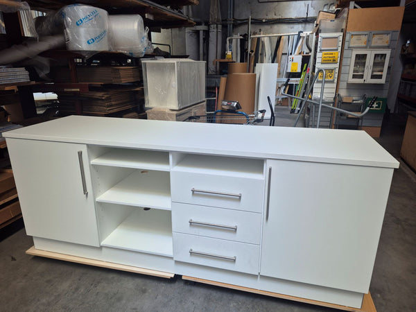 A white modern retail storage cabinet with three drawers, two cabinet doors, and open shelving, photographed in a warehouse setting.