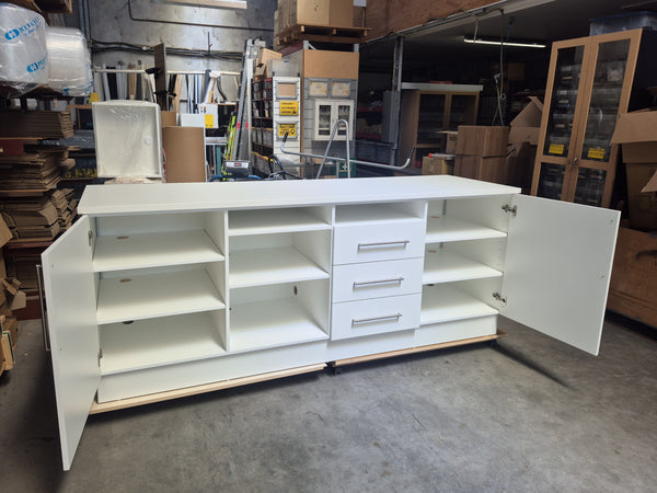 A white modern retail storage cabinet with three drawers, two cabinet doors, and open shelving, shown with all drawers open photographed in a warehouse setting.