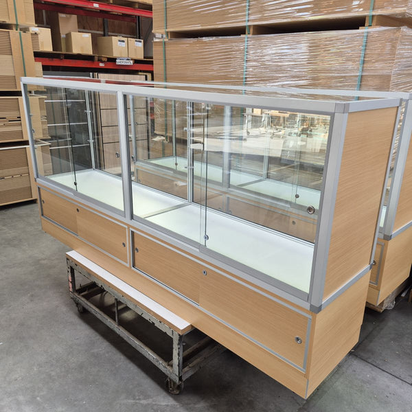 A large commercial glass display case with light wood grain panels and a silver metal frame, shown in a warehouse setting on a metal support cart.