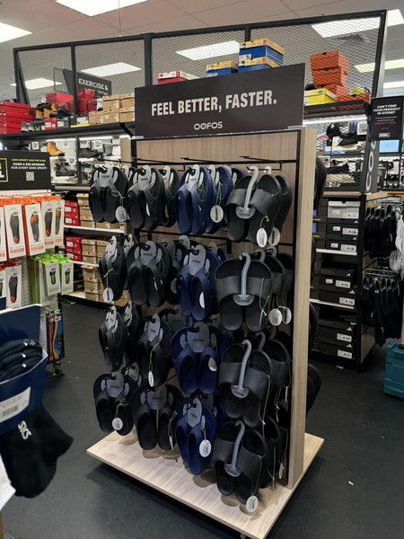 a light oak and black branded OOFOS jandal display stand in a Rebel Sports store