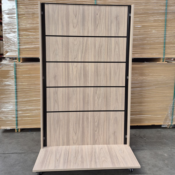 A tall retail display stand with five slatwall inserts featuring a light oak wood grain finish and black metal framing, shown on a warehouse floor with stacked wrapped panels in the background.