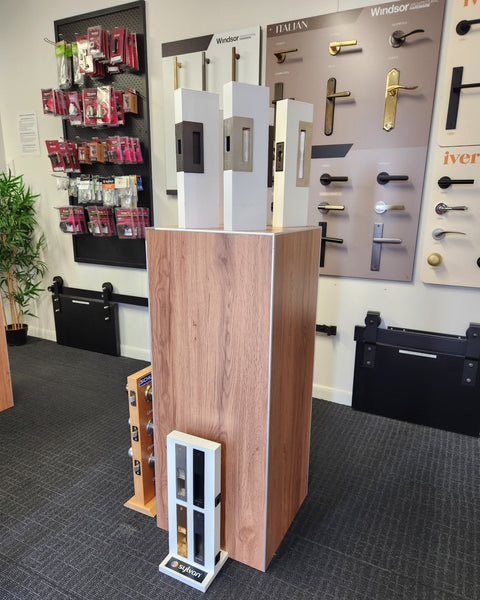 Display of door handles on a free standing floor retail display stand in woodgrain finish and a wooden cabinet in a showroom setting.