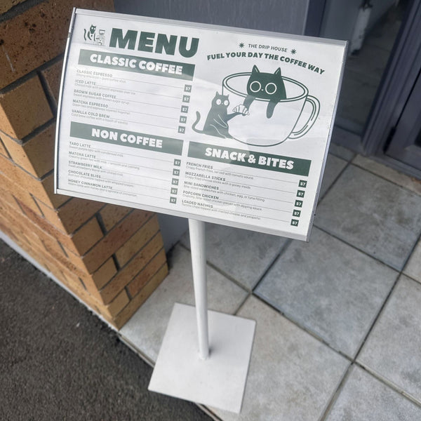 Menu sign for a coffee shop with a cat-themed design on a stand outside a building.