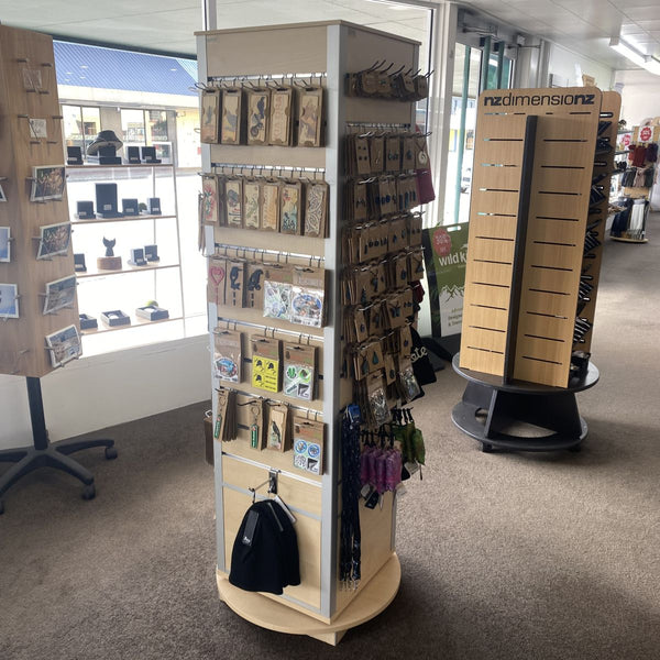 A rotating wooden retail display stand inside a store, featuring a round beige and white central slatwall carousel rack filled with small packaged items, alongside two larger tan display stands in the background.