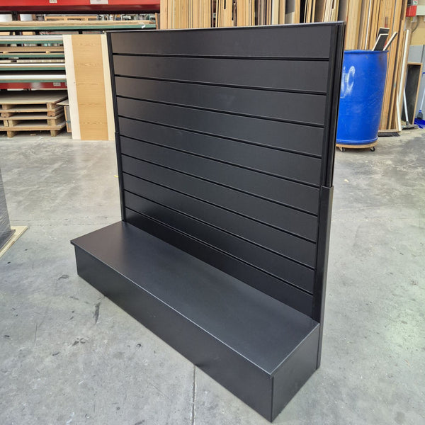 A side view of a black slatwall free standing light weight display stand with a base shelf, photographed in the Plankwall warehouse with concrete floors and wood panels in the background.