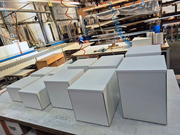 Several plain white rectangular retail floor display plinths arranged on a workbench in a workshop or factory setting with machinery and shelving visible in the background.