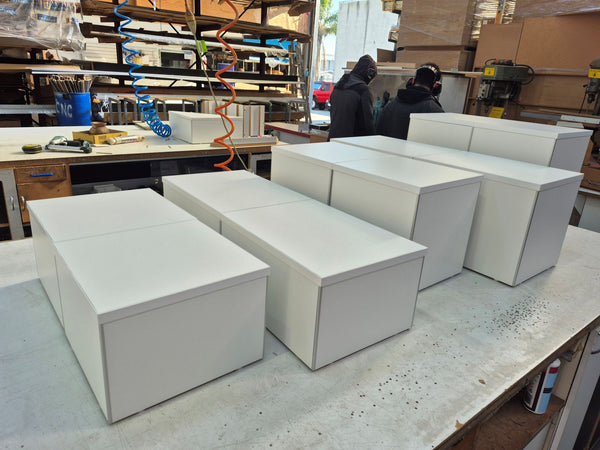 Several white floor display plinths arranged on a workbench in a workshop or studio setting, with shelving and tools visible in the background and two people standing further back.